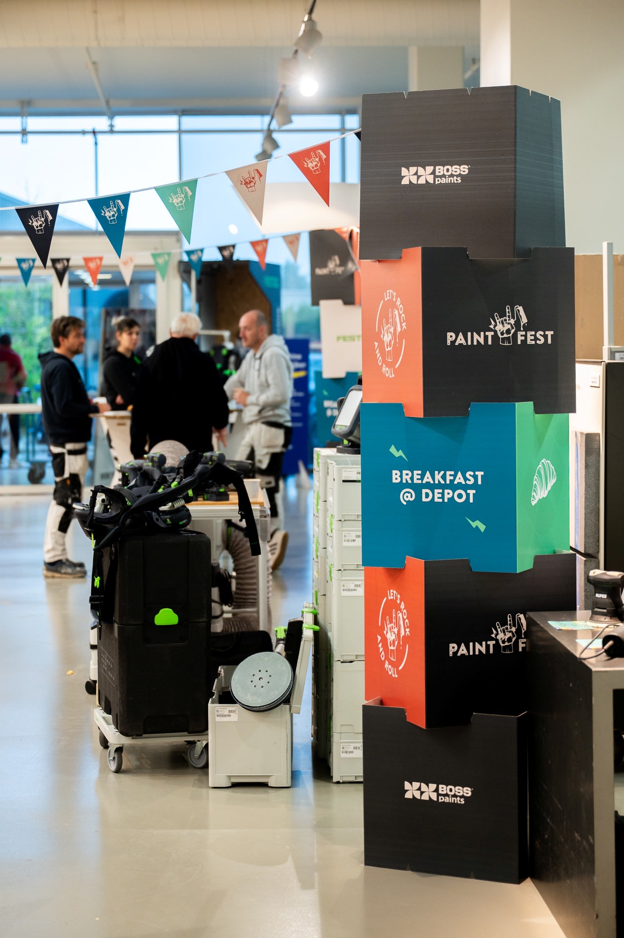 Mensen verzamelen zich binnen bij een bord voor "Paint Fest" en "Breakfast @ Depot" tijdens een evenement, met apparatuur en decoraties zichtbaar op de achtergrond.