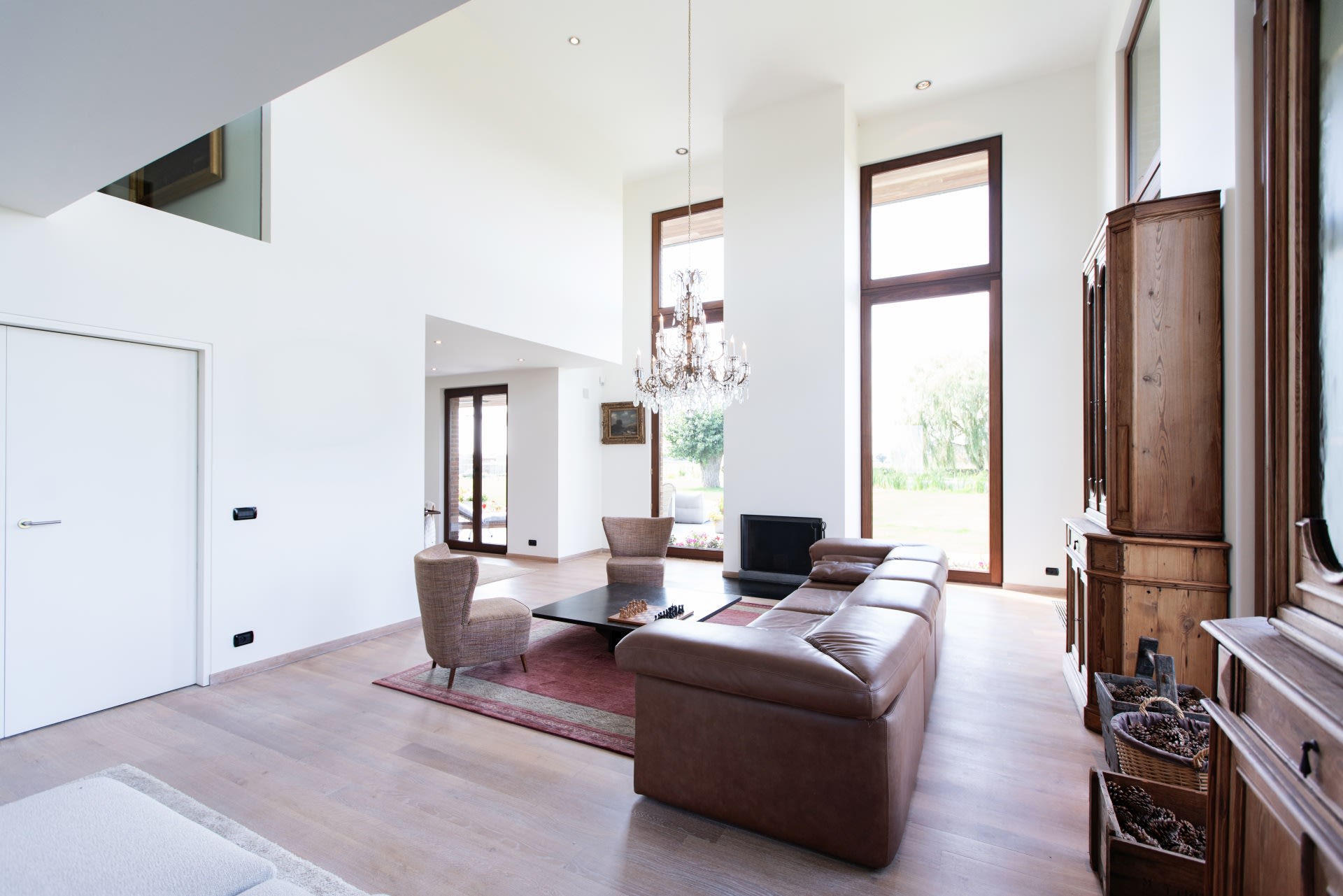Salon moderne avec de hauts plafonds, de grandes fenêtres, un canapé d'angle en cuir marron, des fauteuils, une table basse, un lustre et des meubles en bois, baigné de lumière naturelle.