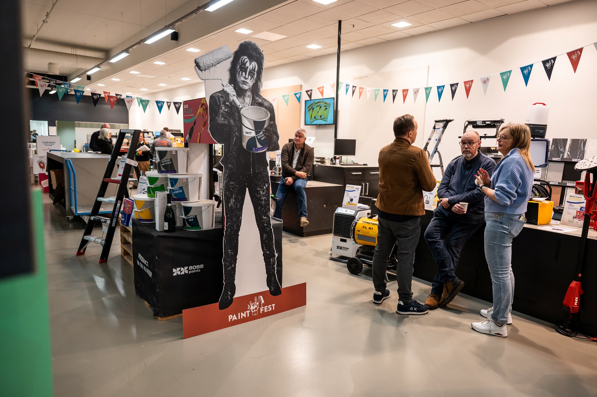 Mensen staan te praten in de buurt van een kartonnen knipsel van een persoon die verfspullen vasthoudt in een versierde binnenruimte met spandoeken en apparatuur.