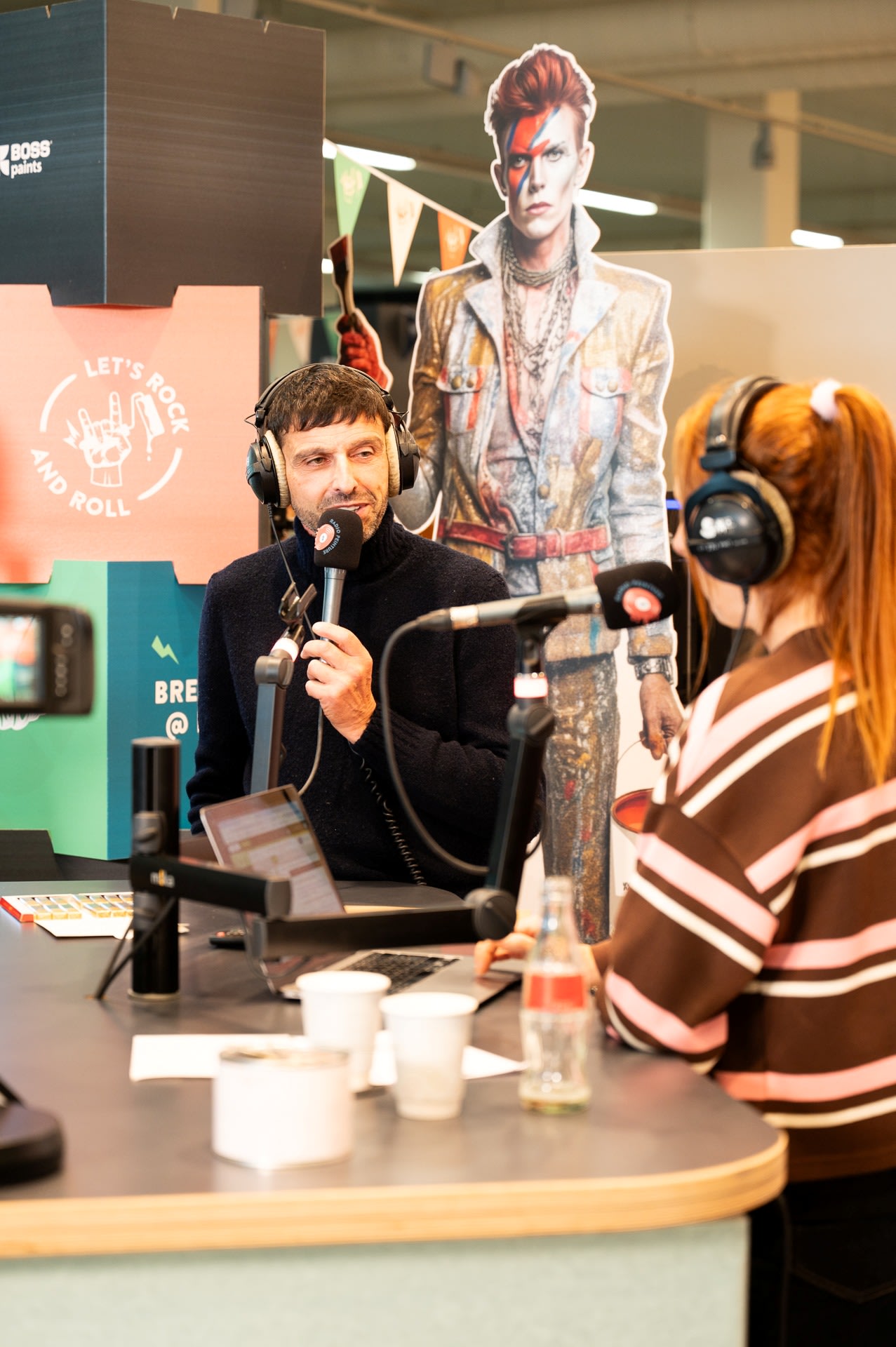 Een man en een vrouw met een koptelefoon op spreken in microfoons bij een radiostation. Een levensgroot kartonnen beeld van David Bowie staat op de achtergrond.