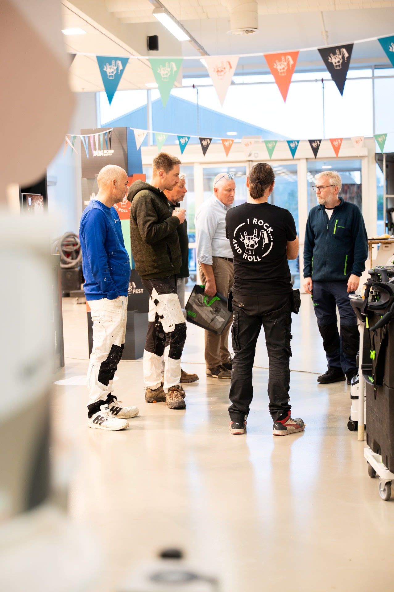 Vijf mensen staan te praten in een helder verlichte binnenruimte versierd met kleurrijke driehoekige vlaggen, met gereedschap en apparatuur zichtbaar op de achtergrond.