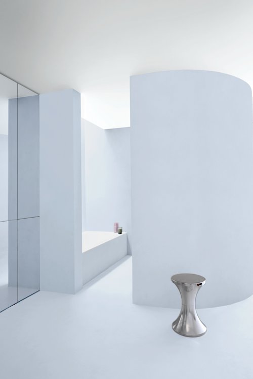 Minimalistische badkamer met gebogen witte muren, een verzonken badkuip, spiegelende oppervlakken en een klein metalen krukje op de voorgrond.