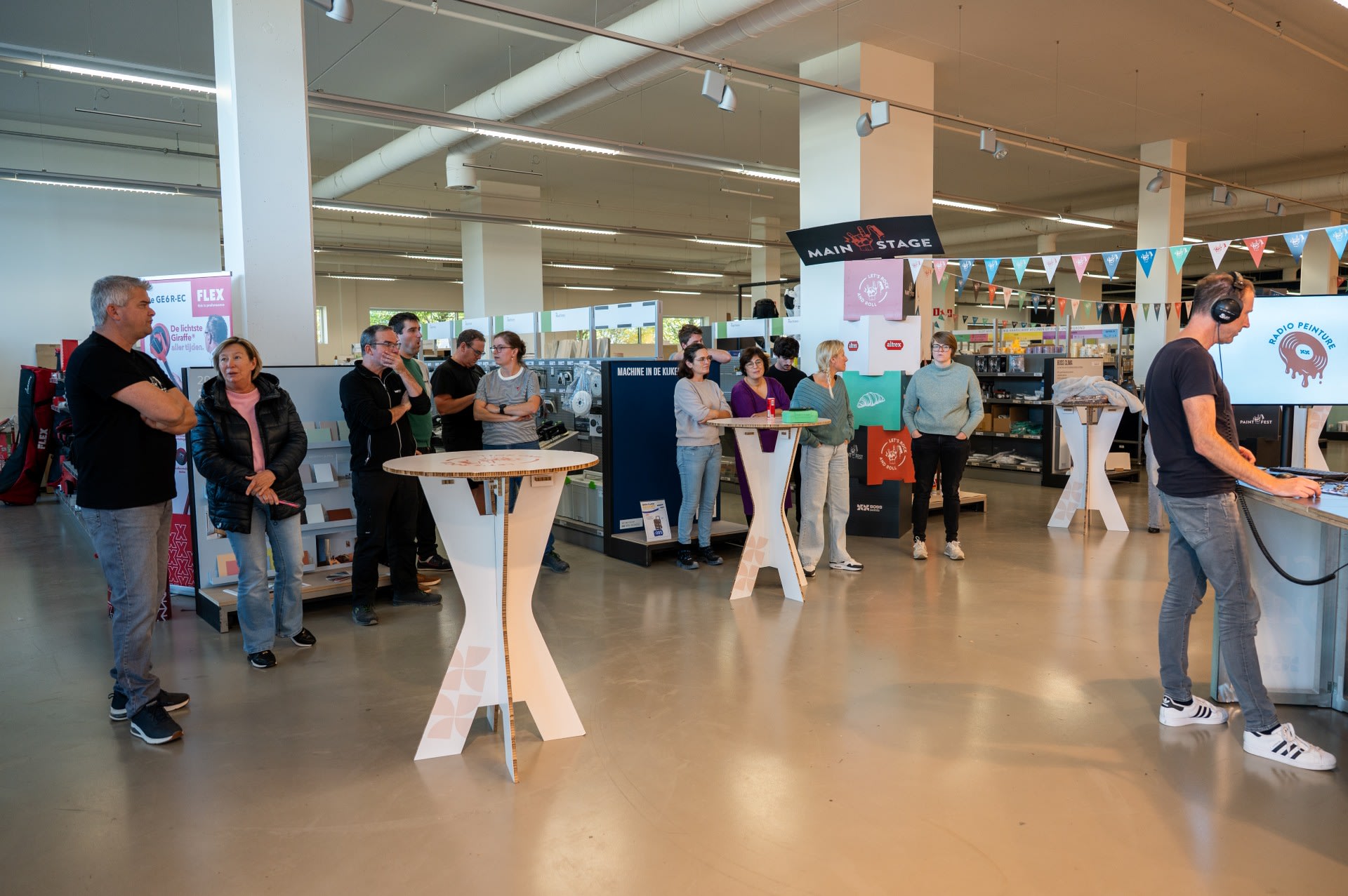 Mensen staan in een winkelachtige omgeving, sommigen bij ronde tafels, terwijl iemand met een koptelefoon op een computer bedient in een kiosk. Slingers versieren het gebied.