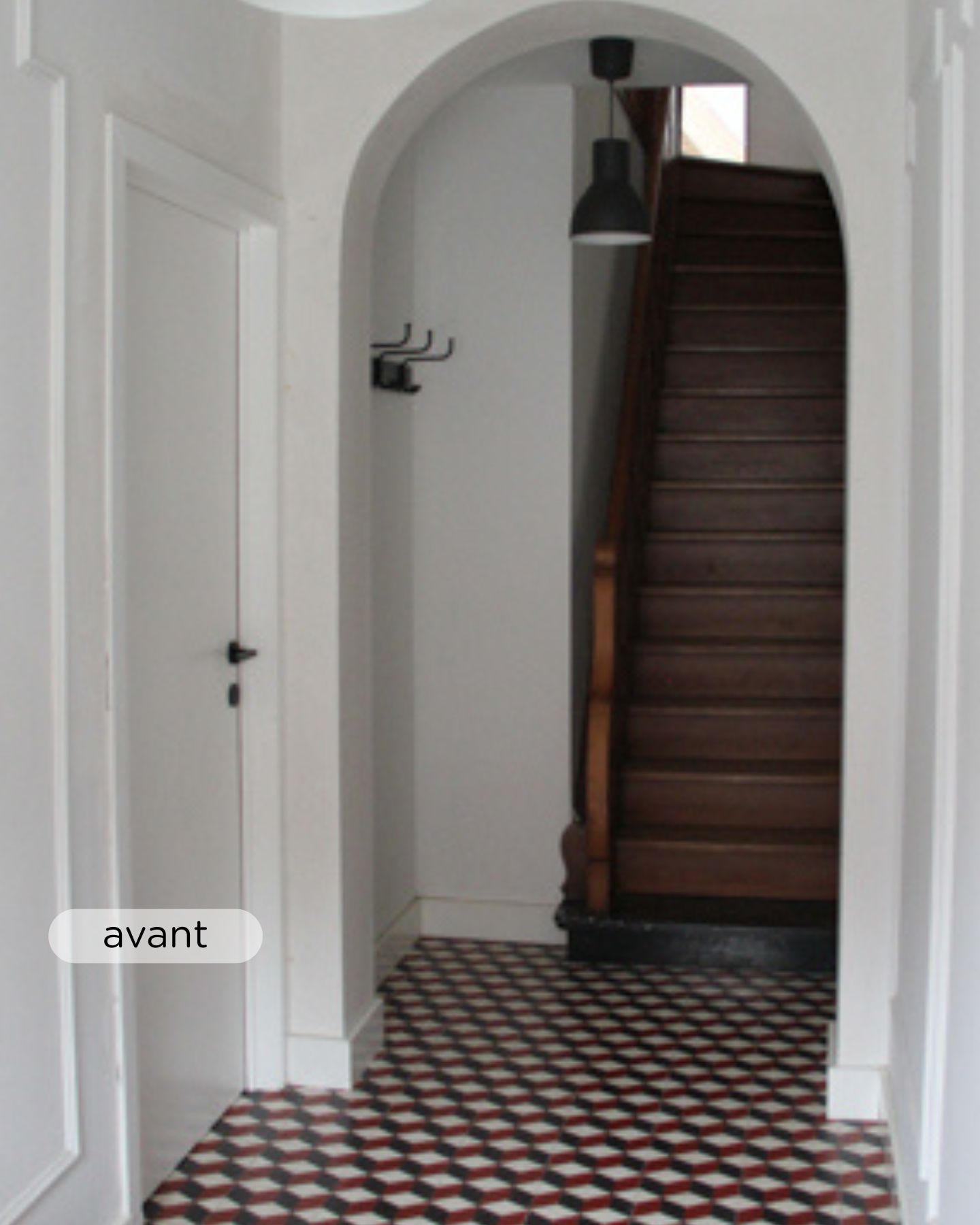 Un couloir étroit avec un sol carrelé à motifs, des murs blancs, une porte blanche fermée sur la gauche, des crochets muraux et un escalier en bois foncé menant à l'étage.