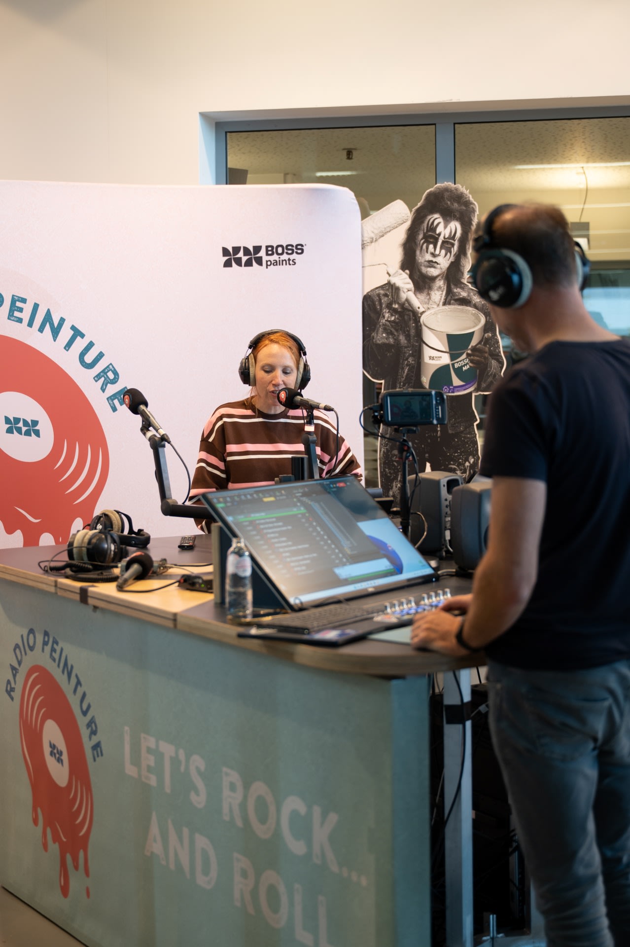 Twee mensen nemen een radioprogramma op bij een zender met de naam "Radio Peinture". De een spreekt in een microfoon terwijl de ander geluidsapparatuur bedient.