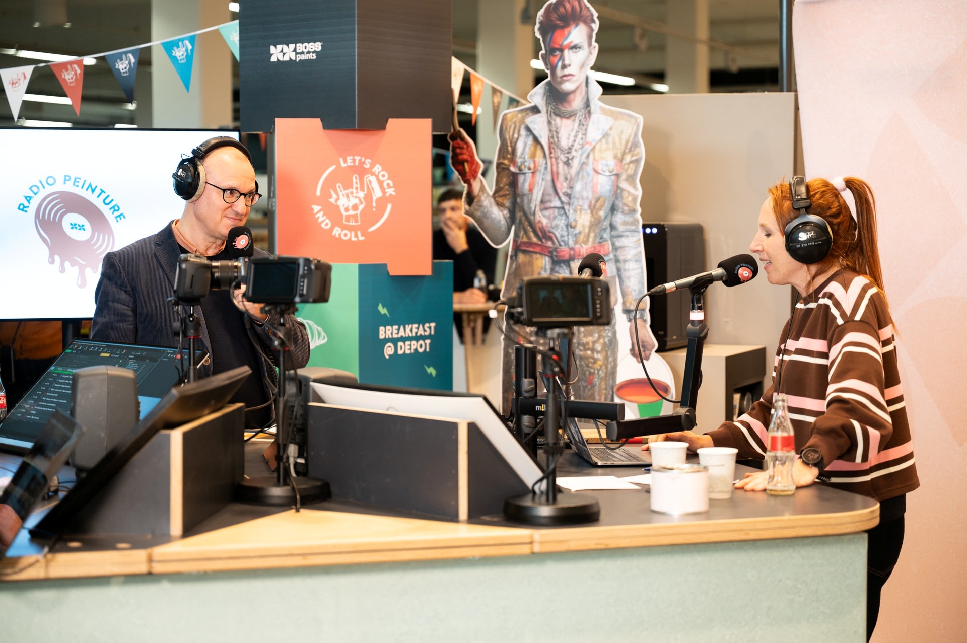 Twee radiopresentatoren zitten aan een bureau met microfoons, opnameapparatuur en een kartonnen knipsel van David Bowie op de achtergrond.