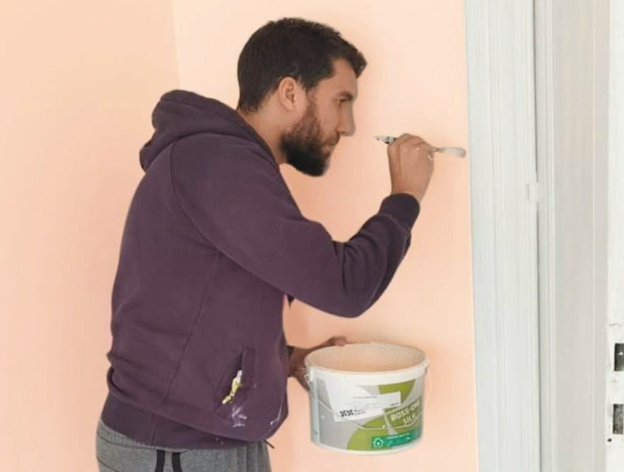 Un homme barbu, coiffé d'une capuche violette, peint délicatement le rebord d'un encadrement de porte blanc à l'aide d'un petit pinceau. Il tient un seau de peinture dans l'autre main et se tient devant un mur couleur pêche.