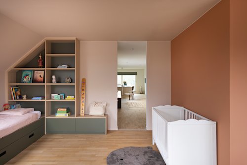 Chambre d'enfant avec un lit encastré, des étagères remplies de jouets et de livres, un berceau blanc et une porte ouverte donnant sur un salon lumineux.