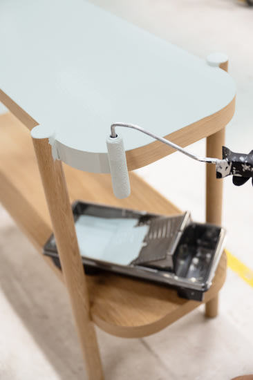 Een persoon gebruikt een verfroller om lichtblauwe verf aan te brengen op de bovenkant van een houten tafel, met een verfbak zichtbaar in de buurt.