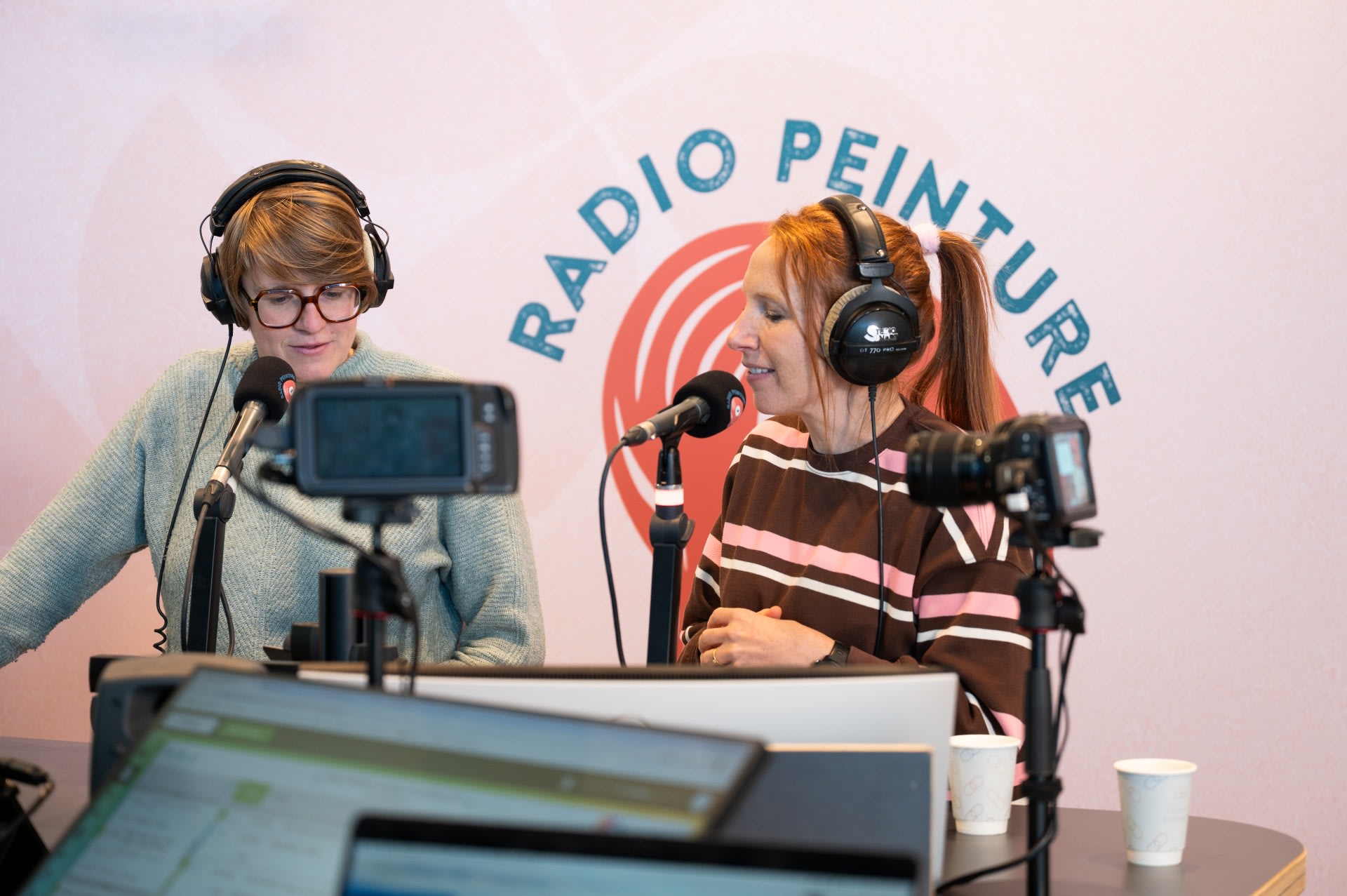 Twee vrouwen met koptelefoons spreken in microfoons in een radiostudio met het label "Radio Peinture. Opnameapparatuur en laptops zijn zichtbaar op de tafel voor hen.