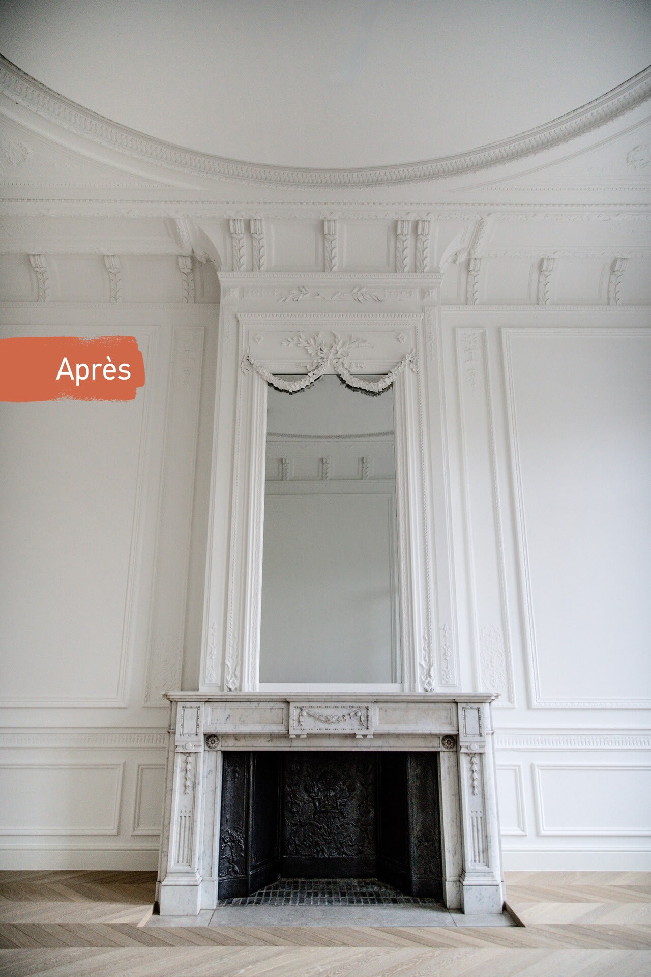 Une cheminée blanche ornée, surmontée d'un grand miroir, se dresse contre des murs aux lambris raffinés, dans une pièce spacieuse et lumineuse. Un échantillon de peinture portant l'inscription « Na » est accroché au mur de gauche.