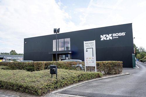 Un bâtiment moderne noir avec un logo « BOSS paints » et une enseigne pour les machines se trouve à côté d’un parking bordé de haies sous un ciel partiellement nuageux.
