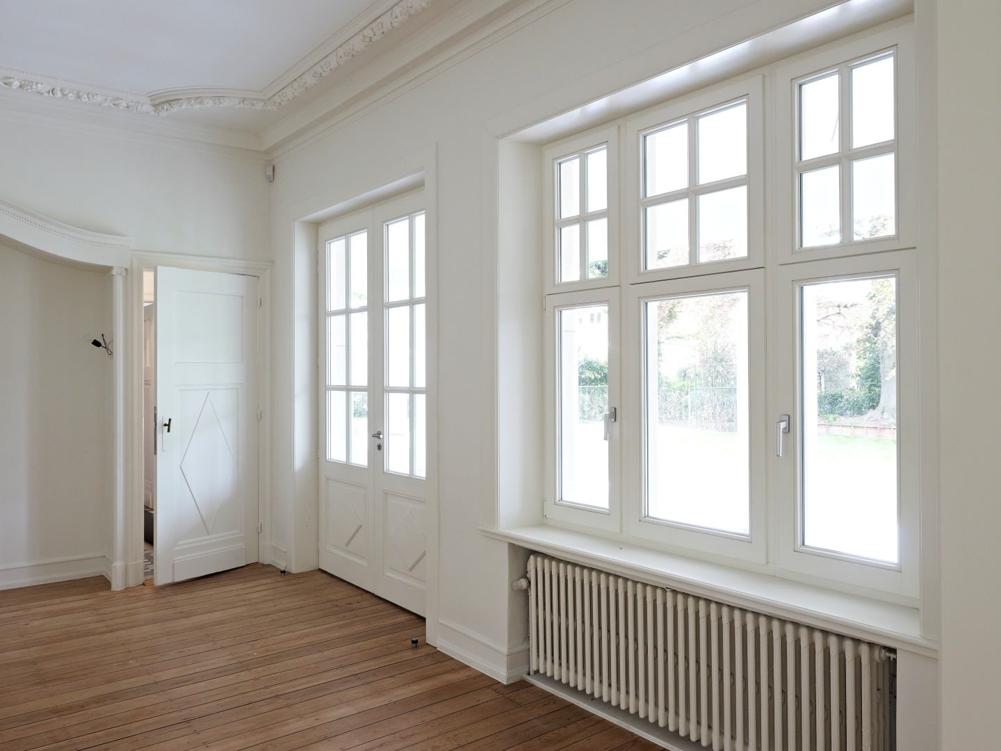 Lege kamer met witte muren, grote ramen, een radiator, houten vloeren en decoratieve kroonlijst op het plafond.