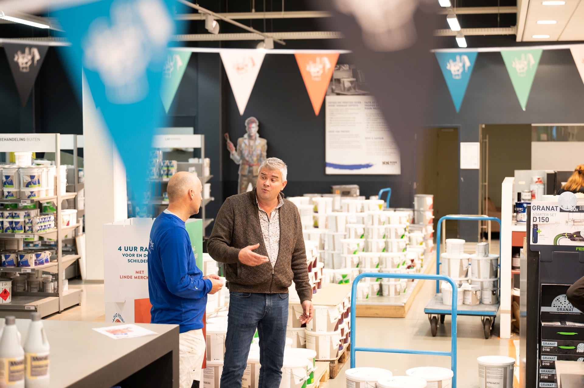 Twee mannen staan te praten in een winkelpad vol met verfblikken en benodigdheden, met informatieborden en vlaggen boven hun hoofd.