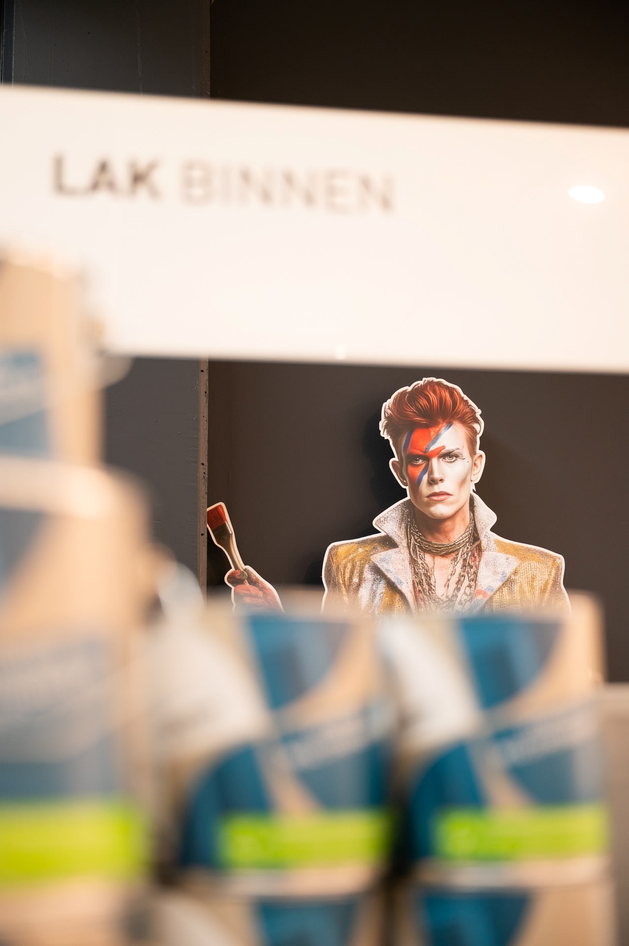 Een kartonnen knipsel van een persoon met een verfkwast staat achter gestapelde verfblikken, met daarboven een bord met de tekst "LAK BINNEN.