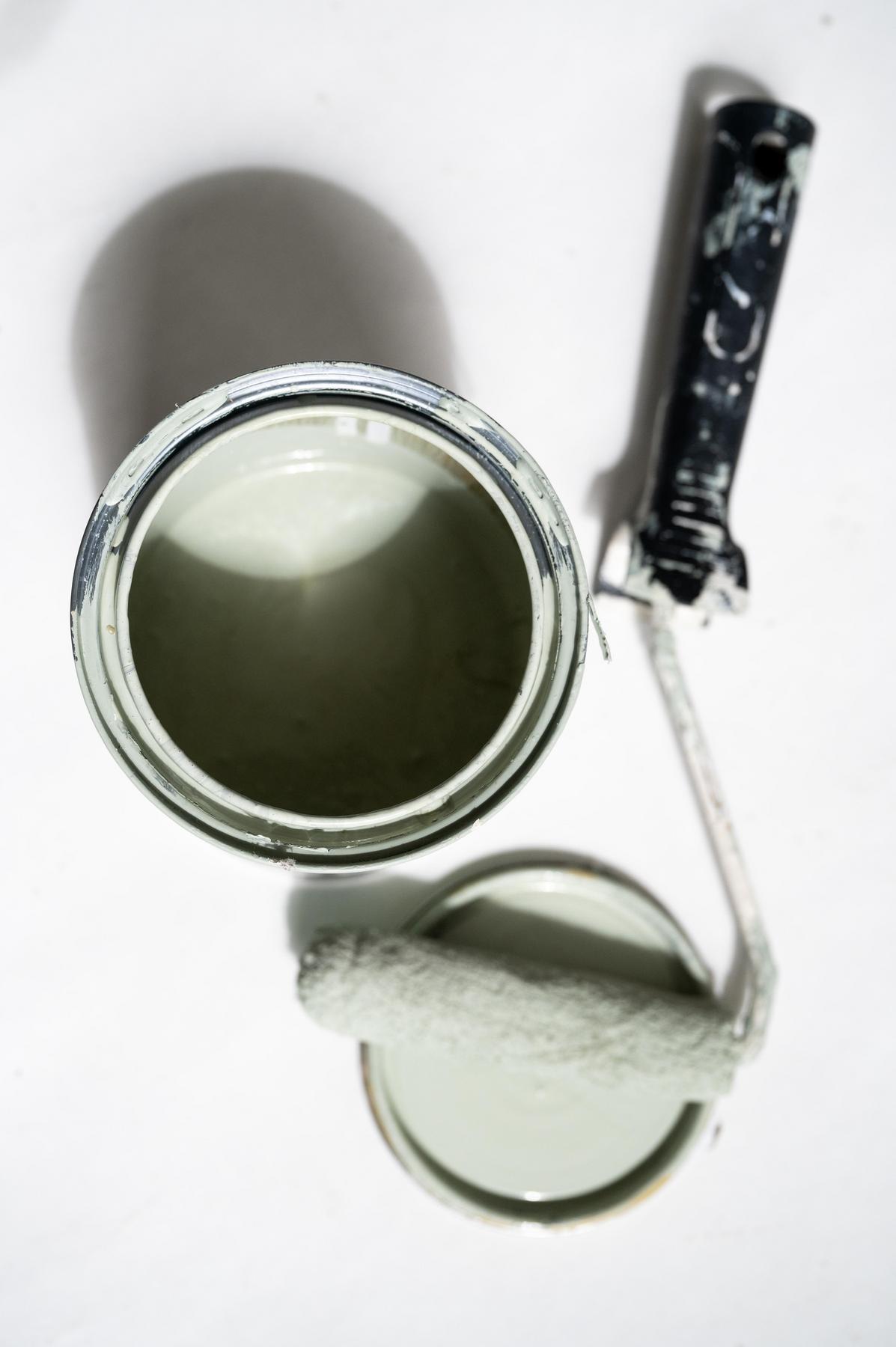 Un pot de peinture vert clair ouvert repose sur une surface blanche, avec son couvercle à côté. Un rouleau à peinture recouvert de la même peinture verte repose sur le couvercle.
