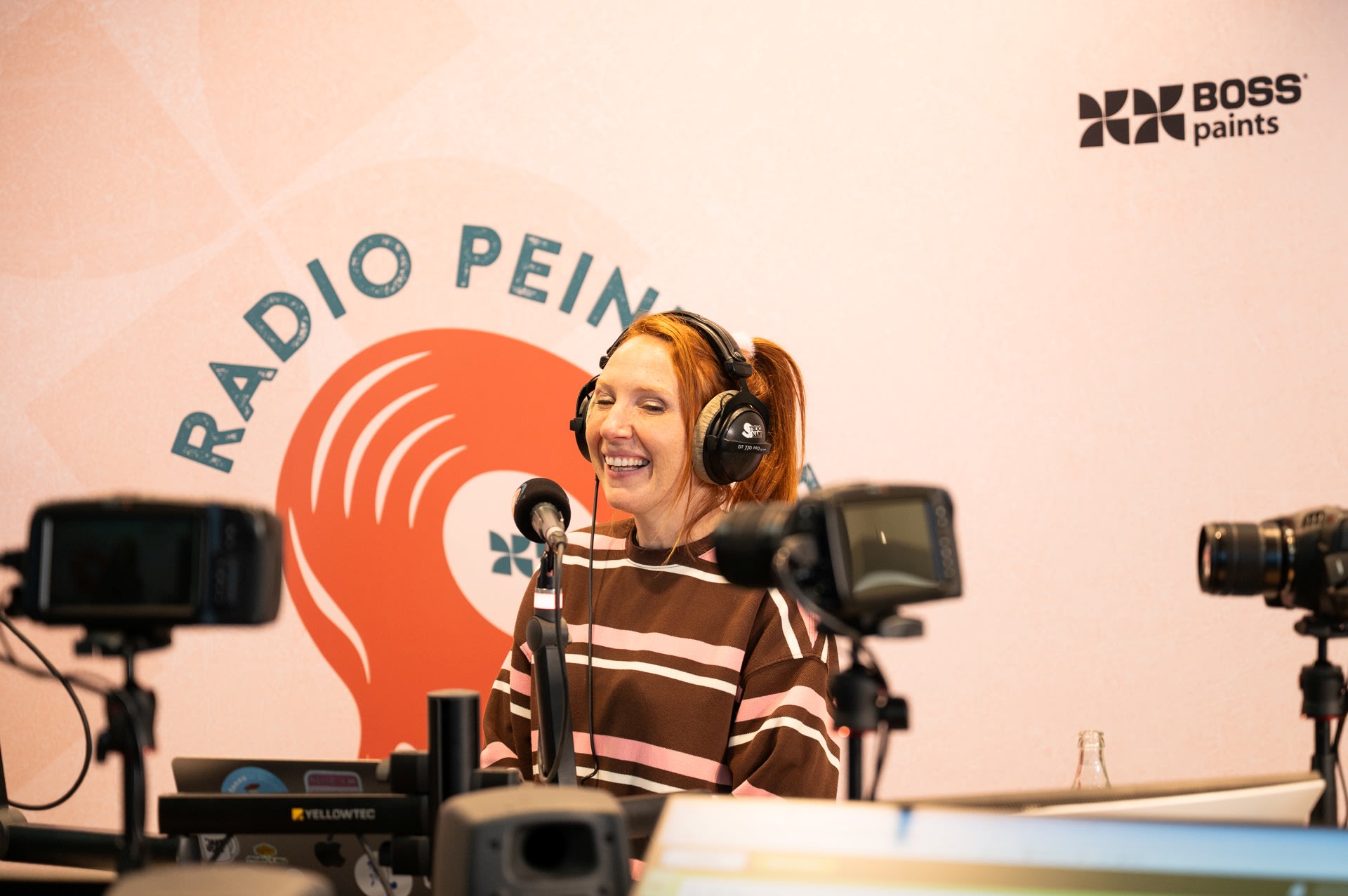 Een vrouw met een koptelefoon op en een gestreept shirt spreekt in een microfoon in een opnamestudio met de naam "Radio Pein" met camera's en apparatuur zichtbaar op de voorgrond.