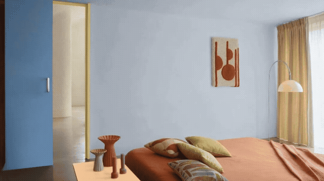 Minimalistische slaapkamer met een bed bedekt met roestkleurig beddengoed en kussens, een lage tafel met vazen, muurkunst, staande lamp, beige gordijnen en een blauwe accentmuur bij de deur.