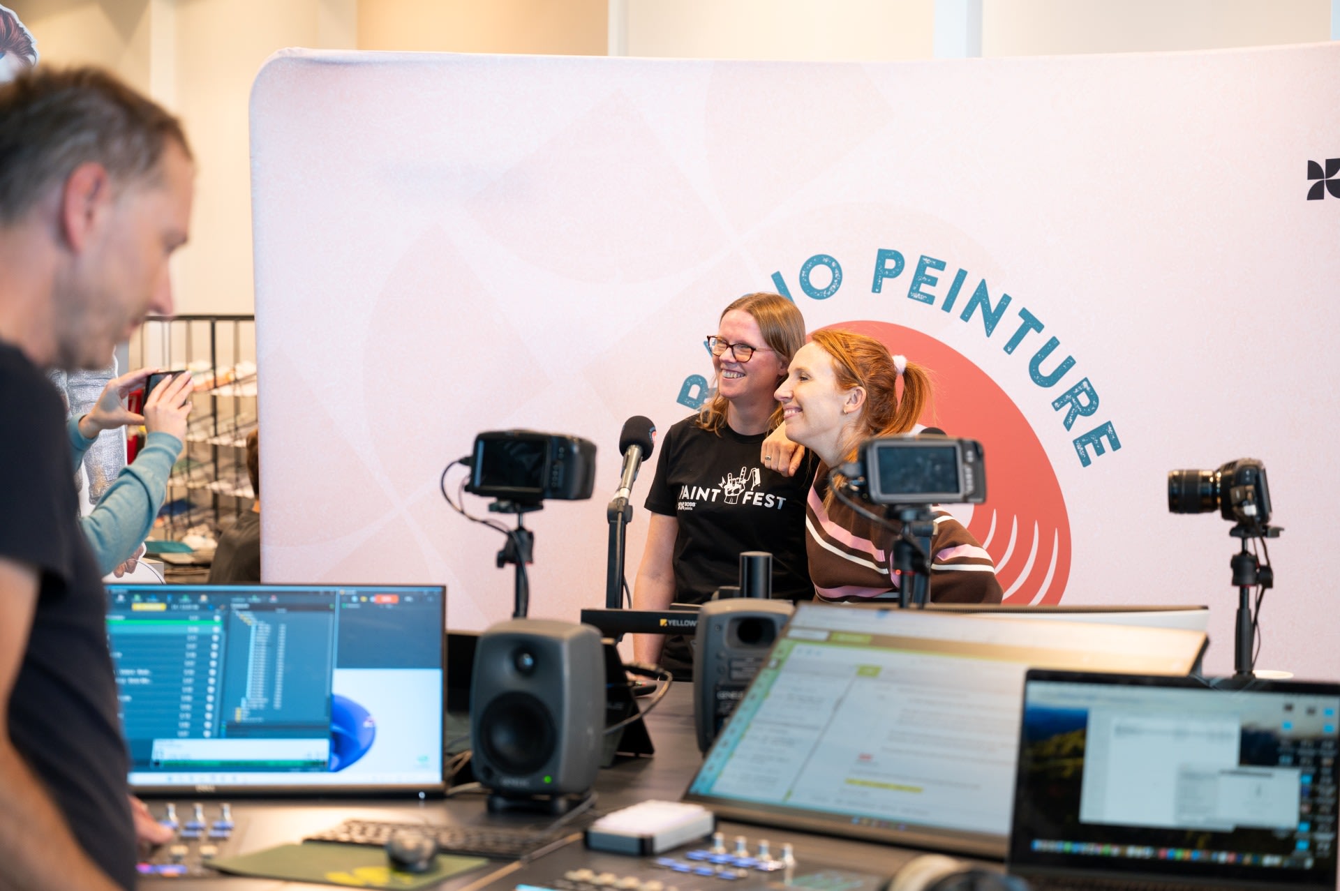 Twee vrouwen staan glimlachend voor een "Radio Peinture" achtergrond terwijl mensen en opnameapparatuur zichtbaar zijn op de voorgrond.