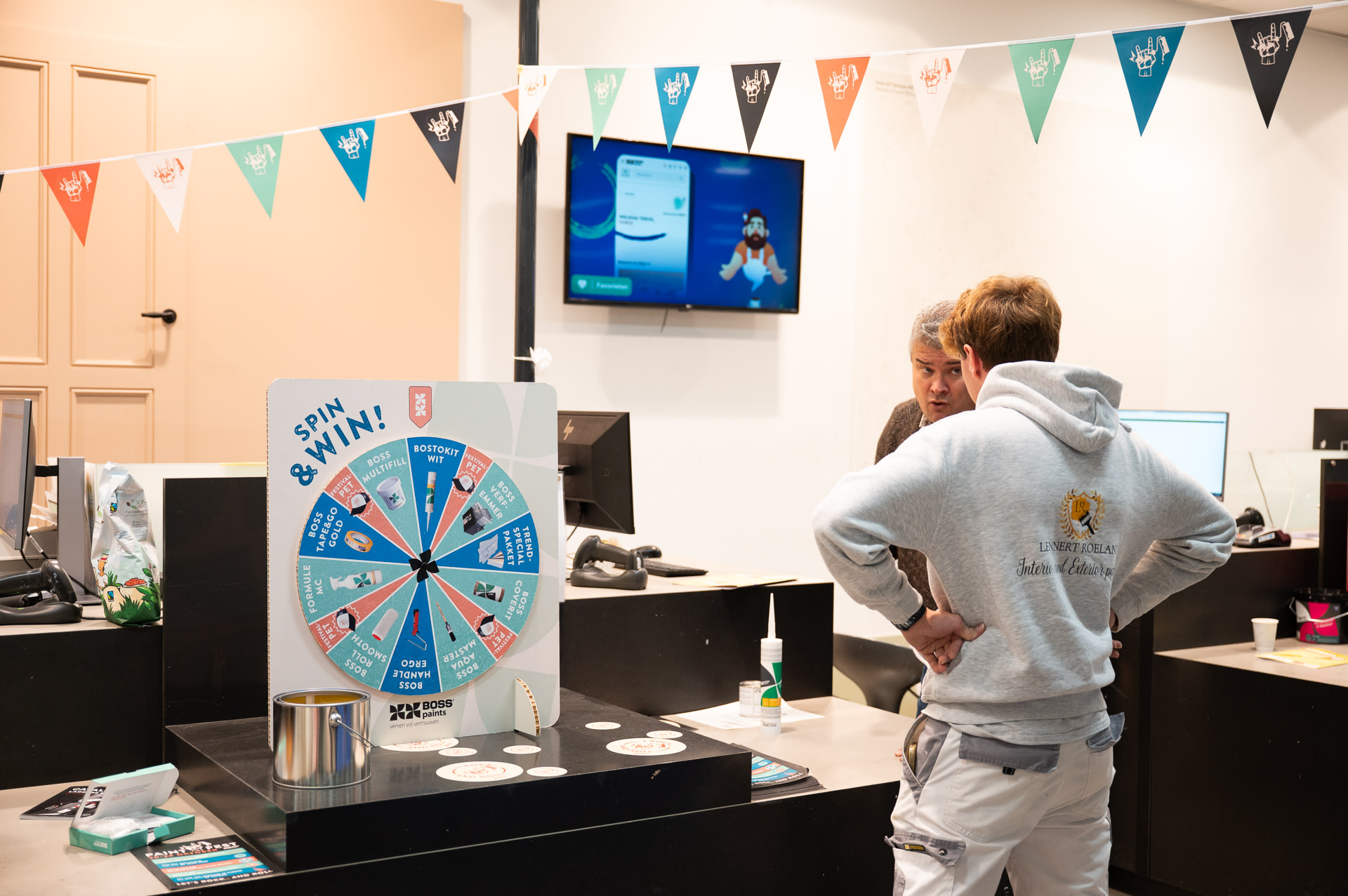 Twee mensen staan bij een toonbank met een "Spin & Win" prijzenrad, banners erboven en een tv-scherm op de achtergrond.