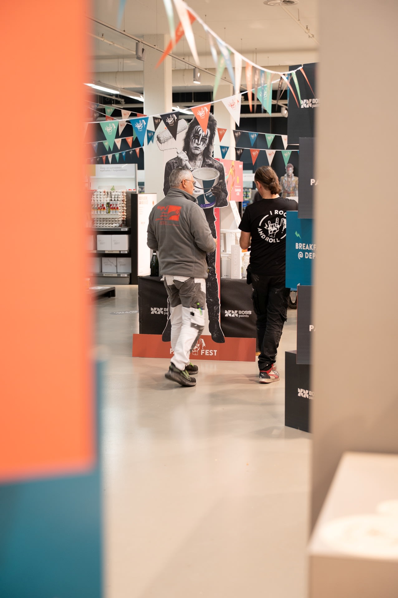 Twee mensen lopen in een versierde binnenruimte met vlaggen en een groot zwart-wit kartonnen knipsel van een persoon.