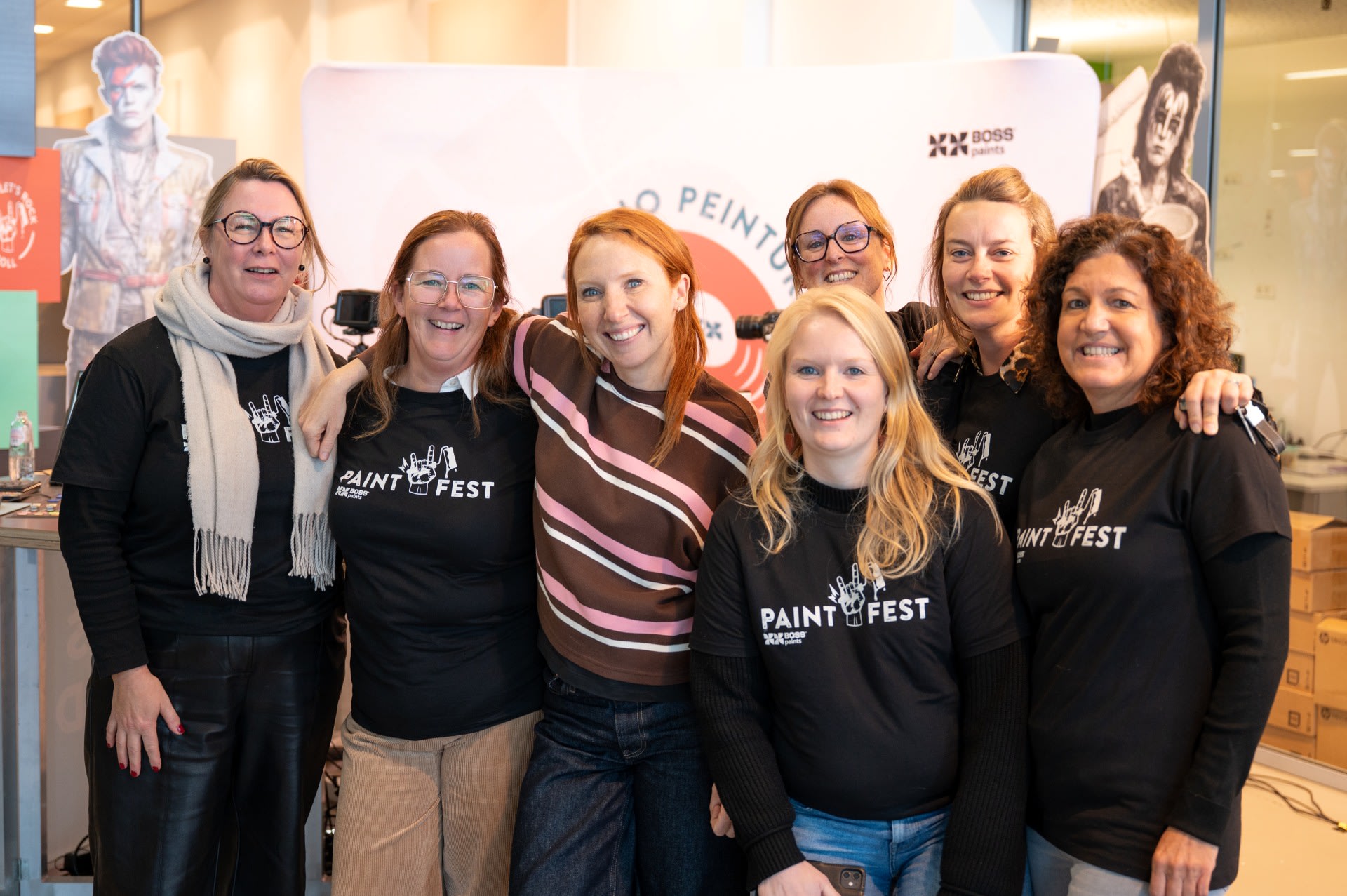 Zeven vrouwen poseren samen binnen, van wie er vier bijpassende zwarte "Paintfest"-shirts dragen. Ze glimlachen en staan voor de achtergrond van een merkevenement.