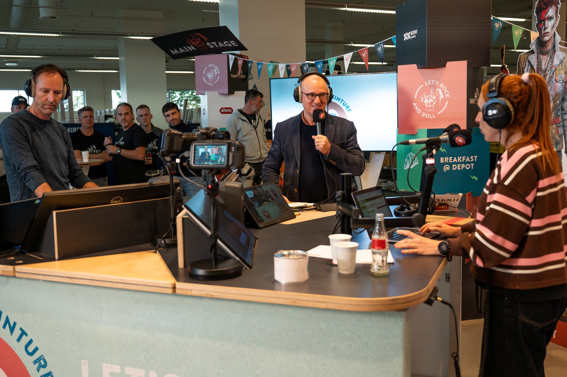 Drie mensen met headsets spreken in microfoons aan een uitzendingstafel, met camera's die opnemen en een publiek dat op de achtergrond staat.