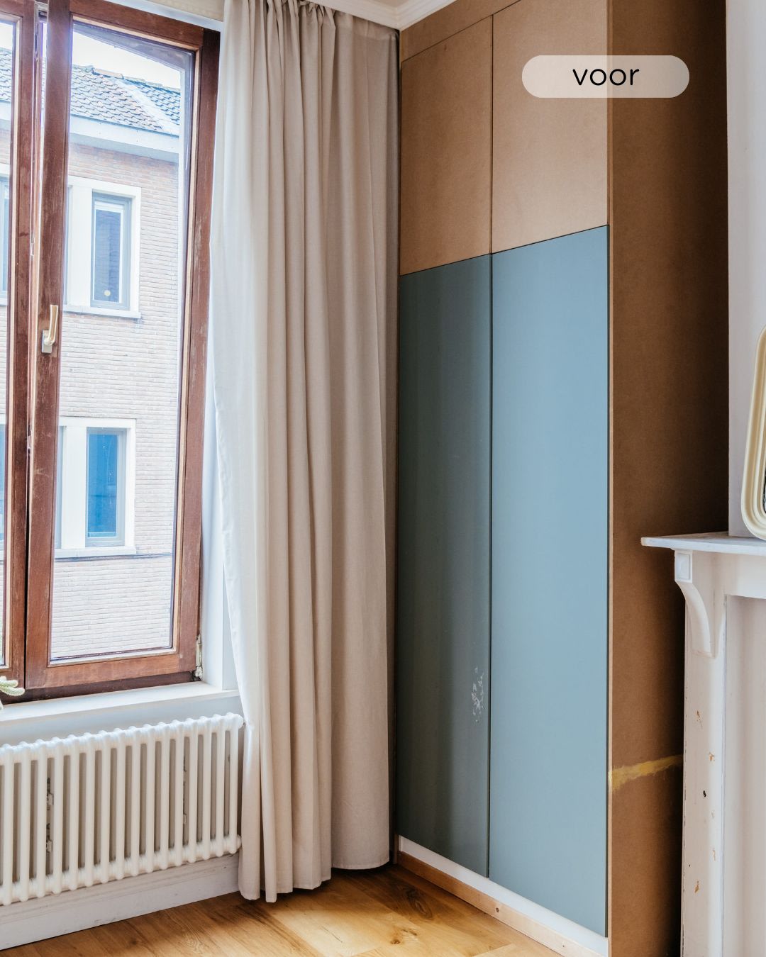 Een hoek van een kamer met een groot raam, beige gordijnen, een radiator, houten vloeren en een kledingkast met blauwe en houten panelen. Het woord "voor" is rechtsboven ingelegd.