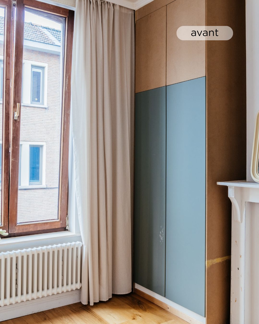Een hoek van een kamer met een groot raam, beige gordijnen, een radiator en een hoge kledingkast met onafgewerkte bruine en blauwe panelen met het label "avant".
