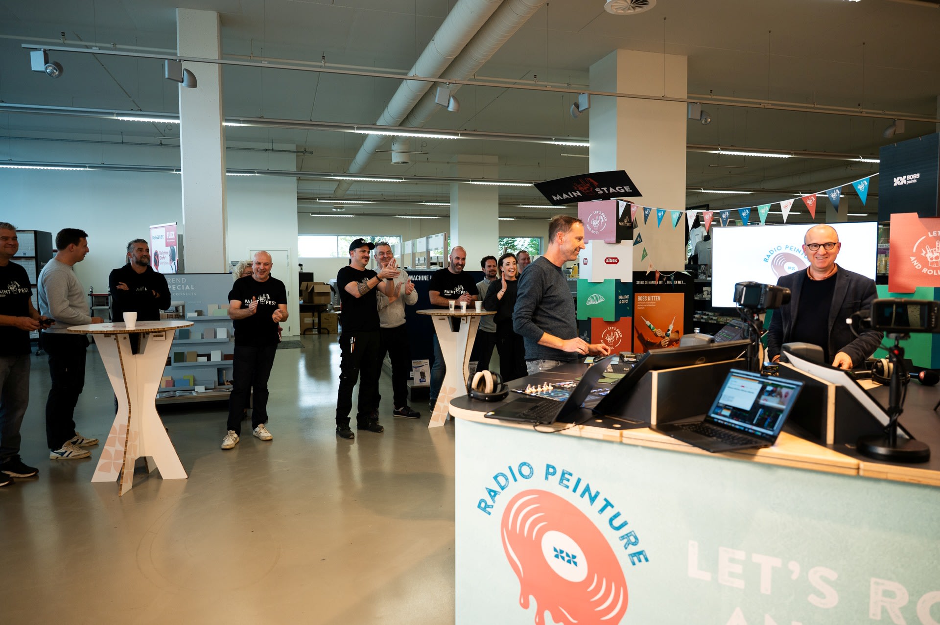 Mensen verzamelen zich in een moderne binnenruimte voor een radio-evenement, met één persoon die spreekt aan een bureau en anderen die eromheen staan, sommigen klappend. Event branding en apparatuur zijn zichtbaar.