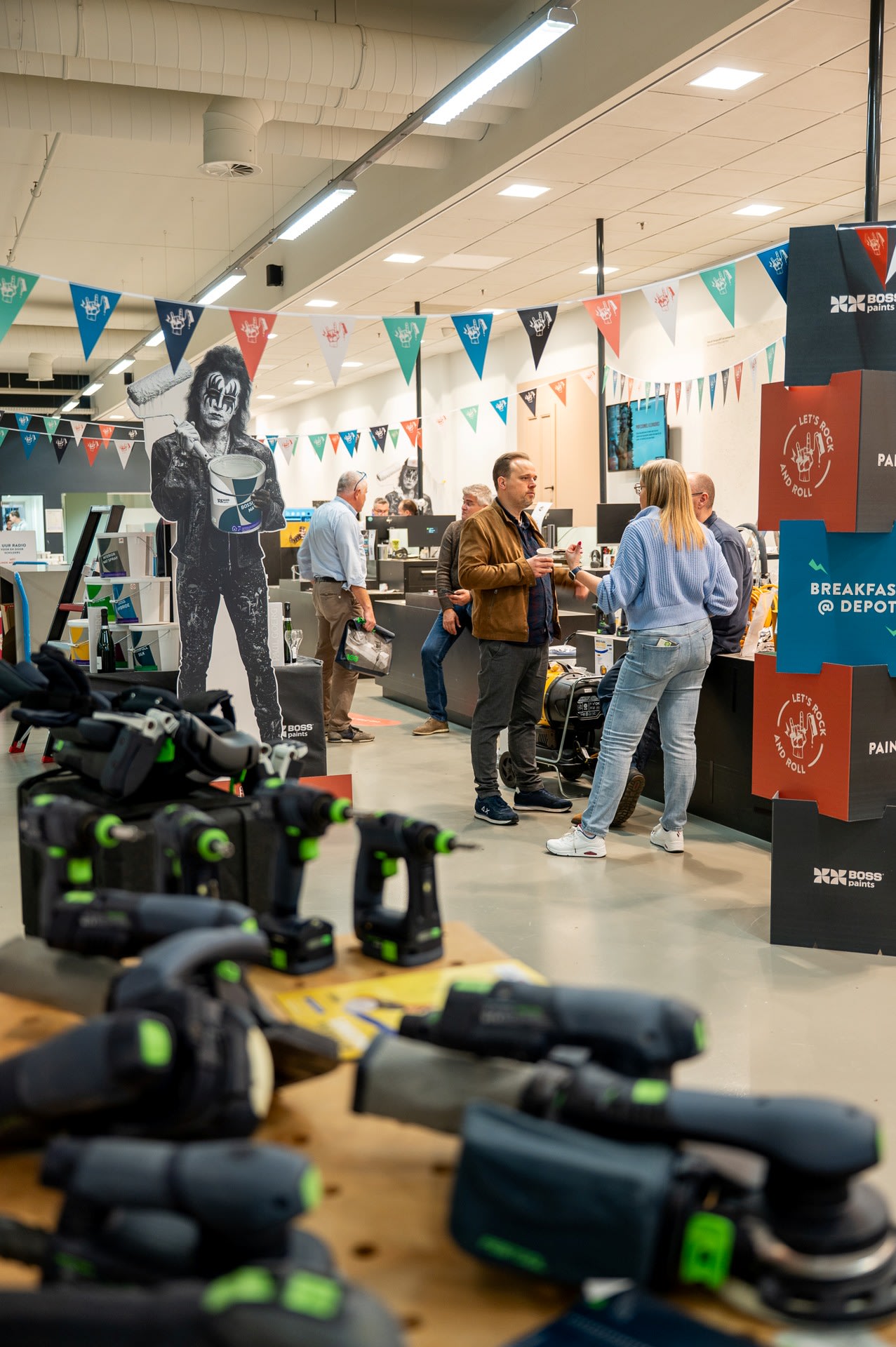 Mensen praten met elkaar in de buurt van een promotiedisplay in een ijzerwinkel, met elektrisch gereedschap op de voorgrond en kleurrijke vlagversieringen erboven.