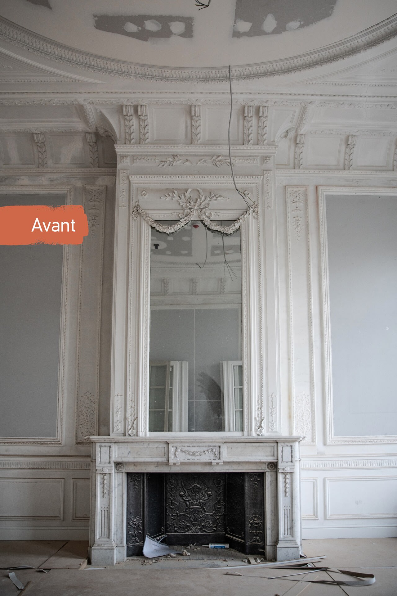Une cheminée blanche ornée, surmontée d'un grand miroir, trône dans une pièce inachevée ; le plafond et les murs présentent des signes de rénovation. Le mot « Voor » figure à gauche.