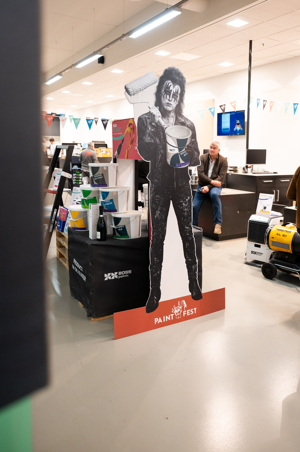 Een levensgroot kartonnen knipsel van een persoon in rockkostuum die verfproducten vasthoudt, staat in een winkel, met rekken verf en mensen op de achtergrond.