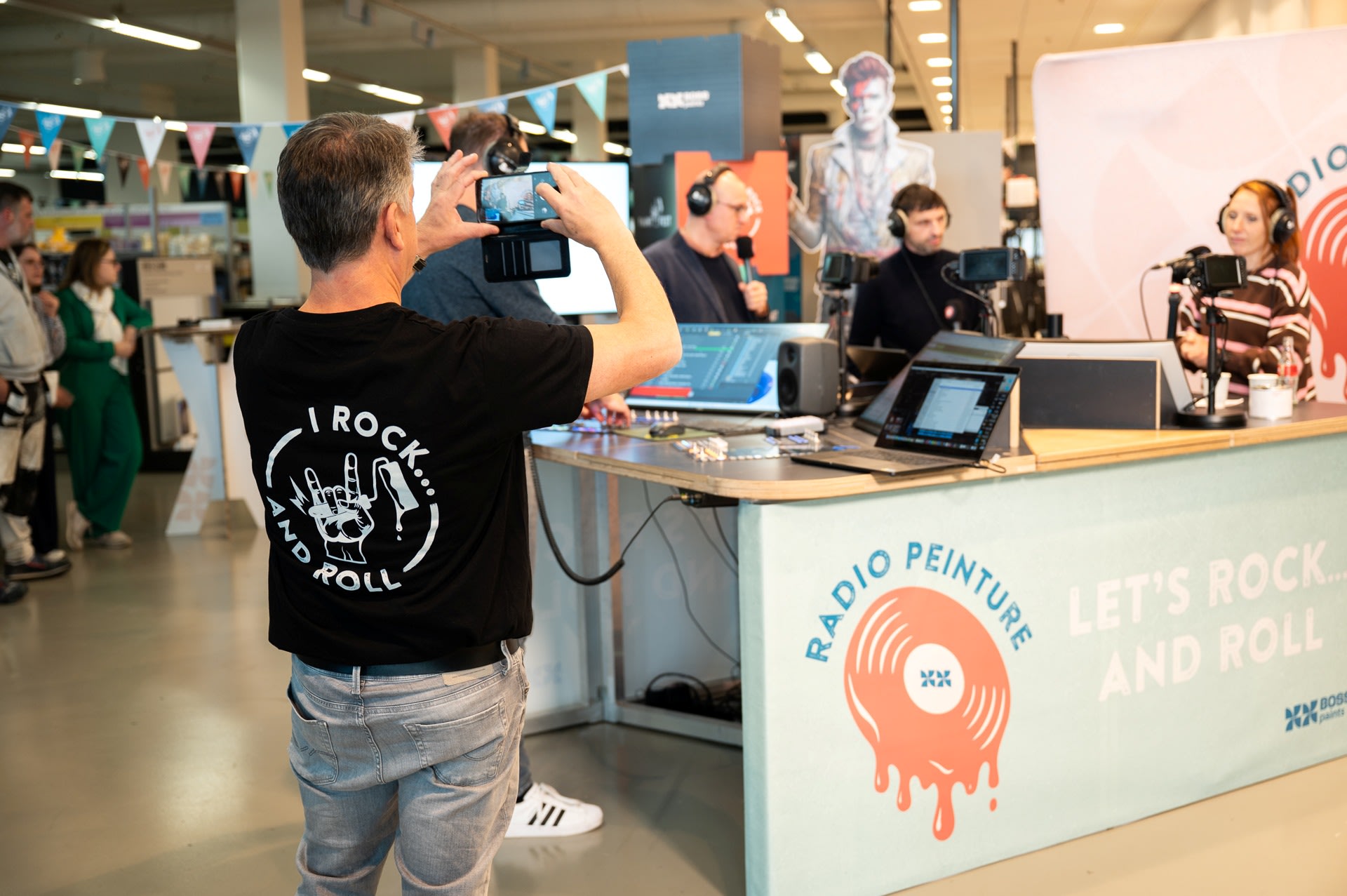 Een man neemt een foto van een live radio-uitzending in een moderne binnenruimte, met presentatoren die spreken in microfoons en apparatuur op tafel.