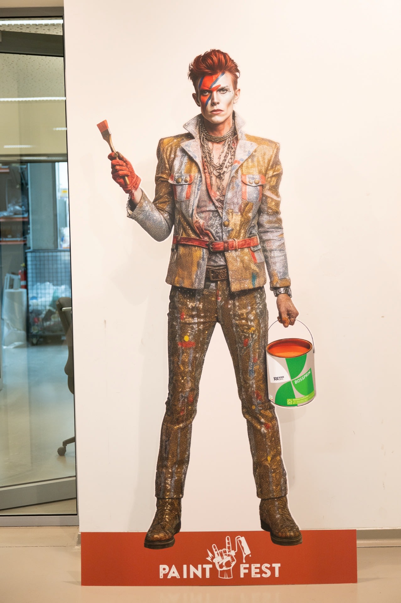 Een levensgroot kartonnen knipsel van een persoon verkleed als Ziggy Stardust van David Bowie, met een verfkwast en een verfblik in de hand, staat voor een muur met een bordje "Paint Fest" aan de voet.