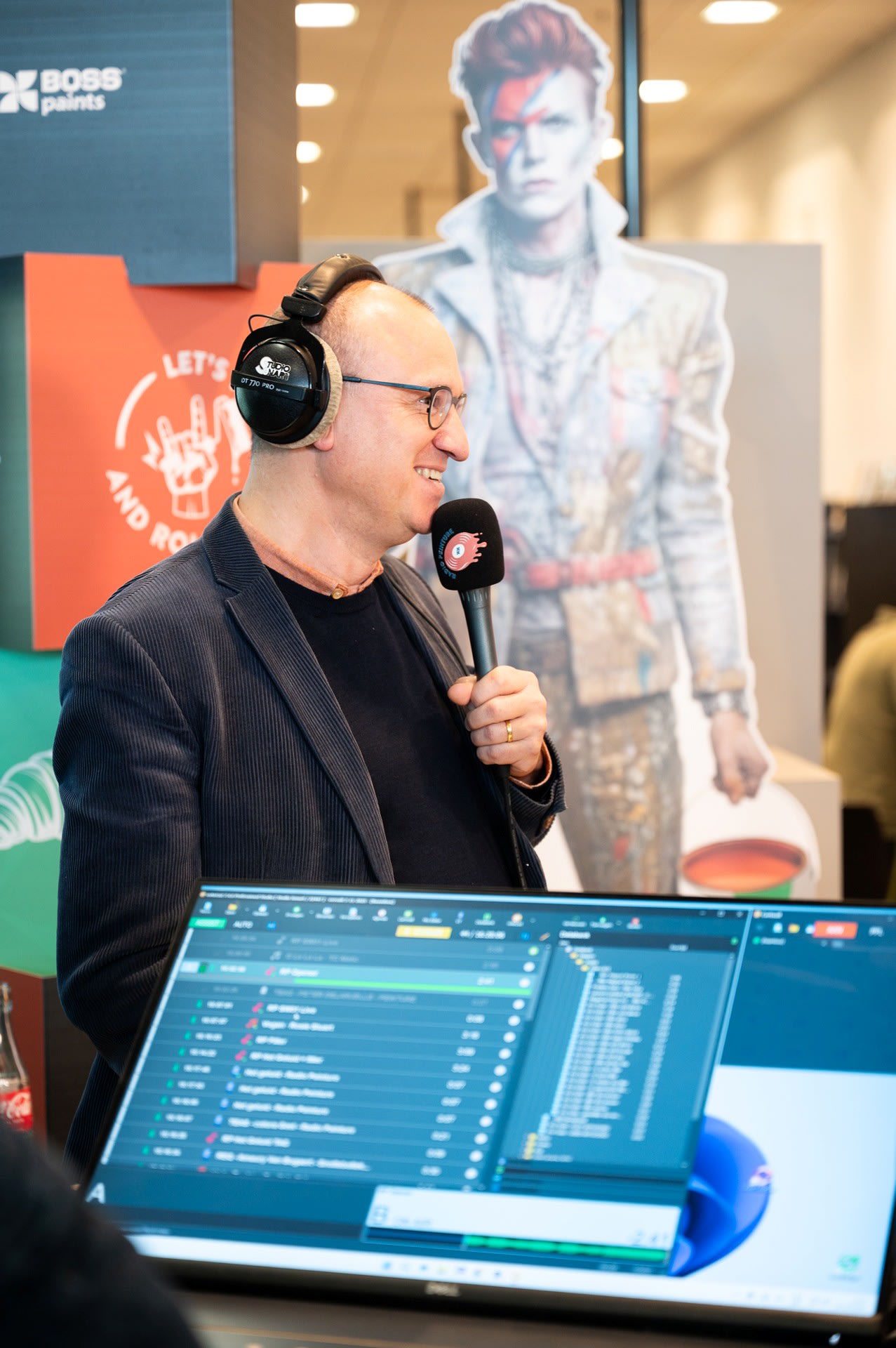 Een man met een koptelefoon op houdt een microfoon vast en spreekt, met een computerscherm op de voorgrond en een David Bowie-knipsel op de achtergrond.