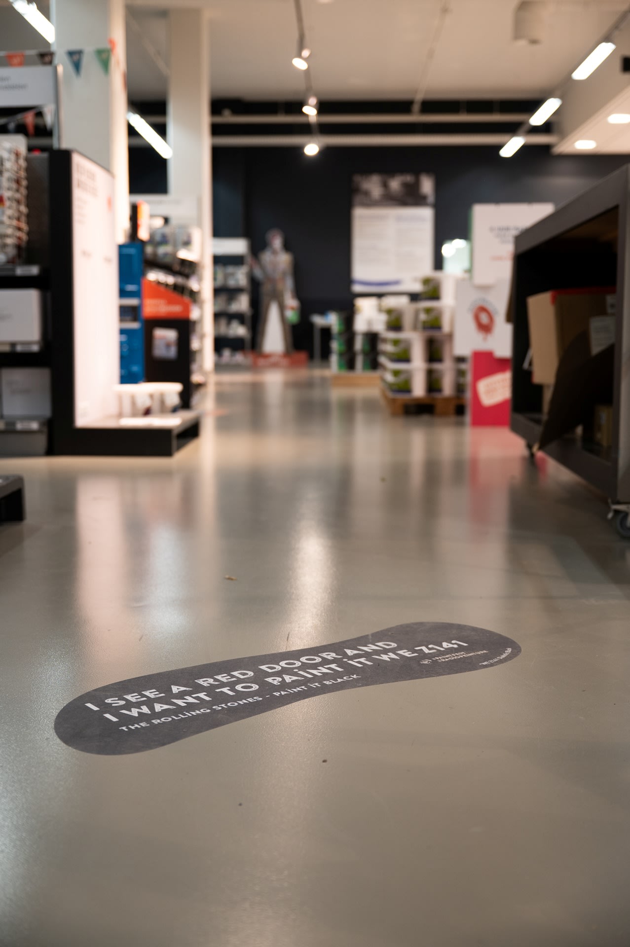 Op een sticker op een winkelvloer staat de tekst: "Ik zie een rode deur en ik wil hem zwart schilderen. The Rolling Stones, Paint It Black, 1966." Op de achtergrond zijn schappen en producten zichtbaar.