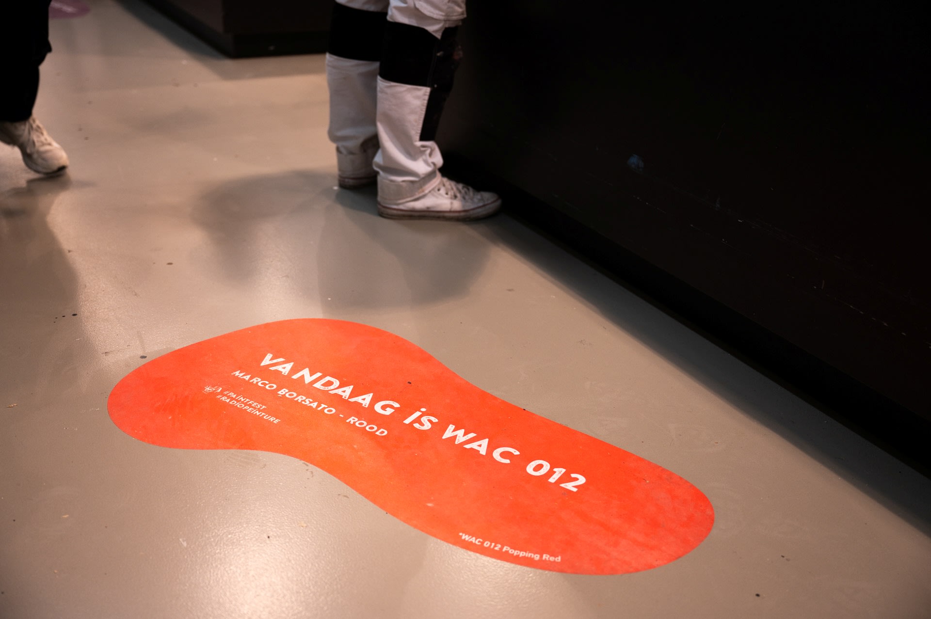 Op een rood bord op de vloer staat "VANDAAG IS WAC 012" in het Nederlands; mensen staan bij een zwarte toonbank, met alleen hun benen zichtbaar.