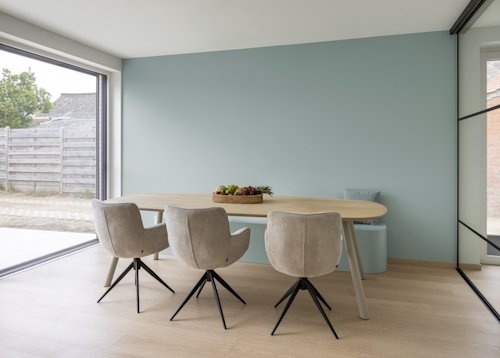Une salle à manger moderne avec une table en bois, trois chaises beiges, un bol de fruits et une œuvre d'art murale sur laquelle est écrit « NOTRE MAISON / NOUVELLE MAISON » en texte noir.