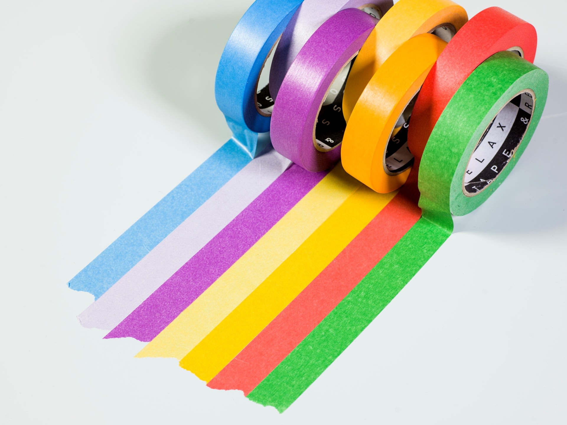 Cinq rouleaux de ruban adhésif coloré – bleu, violet, jaune, orange et vert – sont déroulés en bandes parallèles sur une surface blanche.