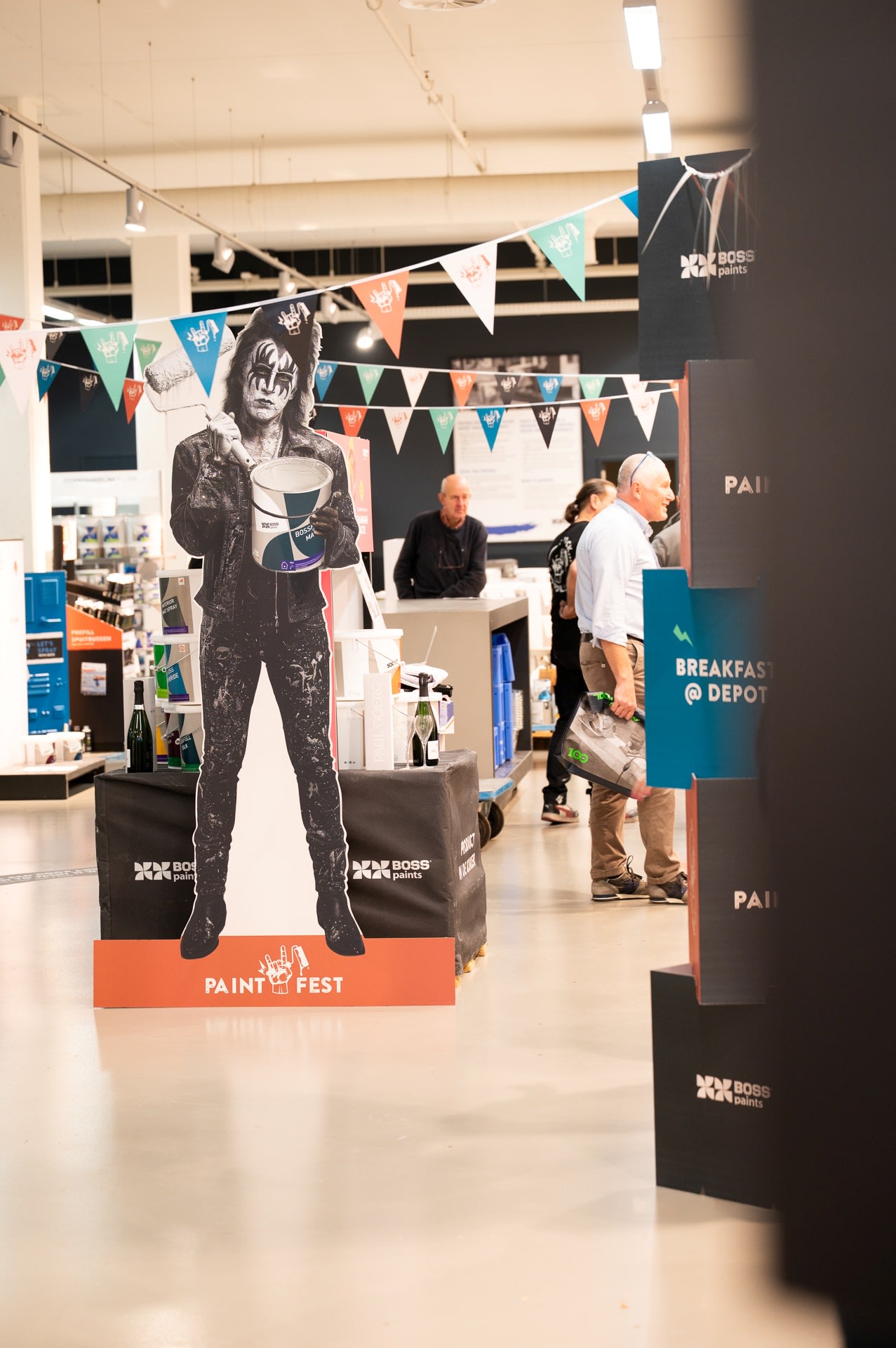 Een kartonnen knipsel van een geschminkt persoon die een verfblik vasthoudt, staat in een winkel in de buurt van "Paint Fest"-borden, met klanten en personeel op de achtergrond.