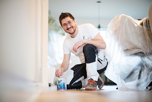 Een lachende man in werkkleding knielt op de grond met een verfkwast naast een open verfblik, met meubels bedekt met plastic op de achtergrond, wat suggereert dat hij een kamer aan het schilderen is.