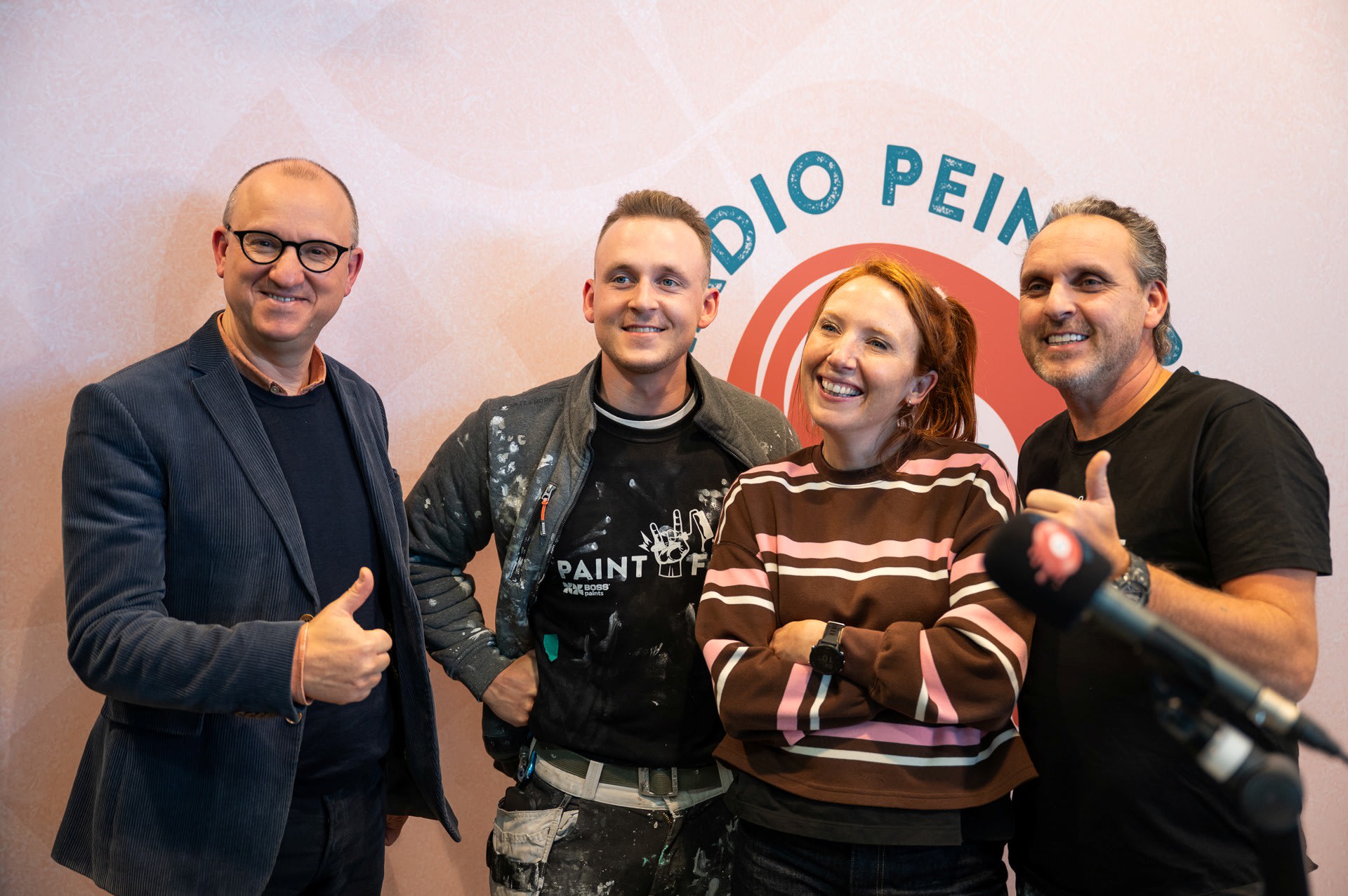 Vier mensen staan voor een muur met "RADIO PEINTURE" gedeeltelijk zichtbaar, glimlachen en geven duimen omhoog, met een microfoon op de voorgrond.