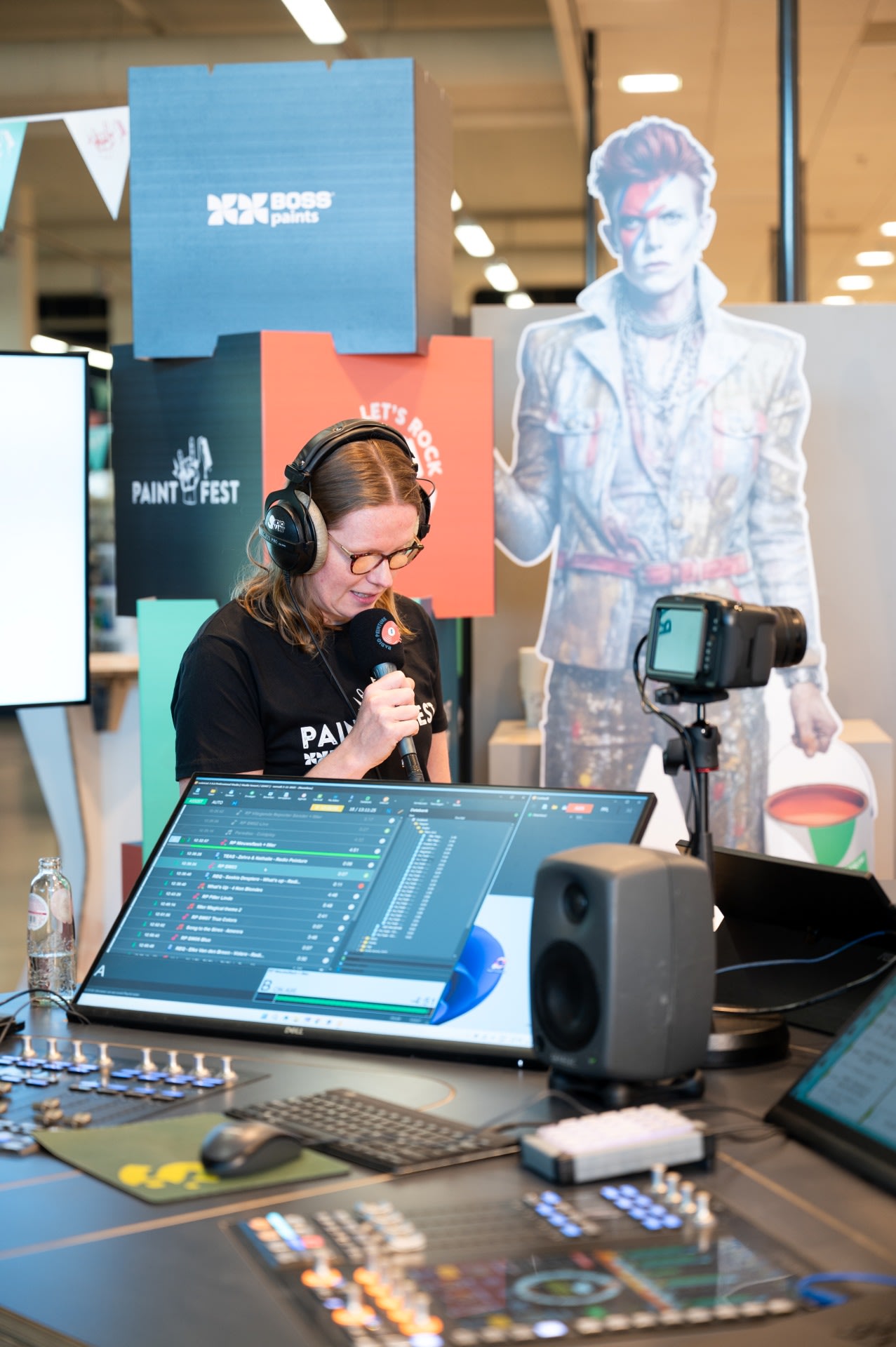 Een persoon met een koptelefoon spreekt in een microfoon op een werkstation met audioapparatuur en computerschermen, met een David Bowie-knipsel op de achtergrond.