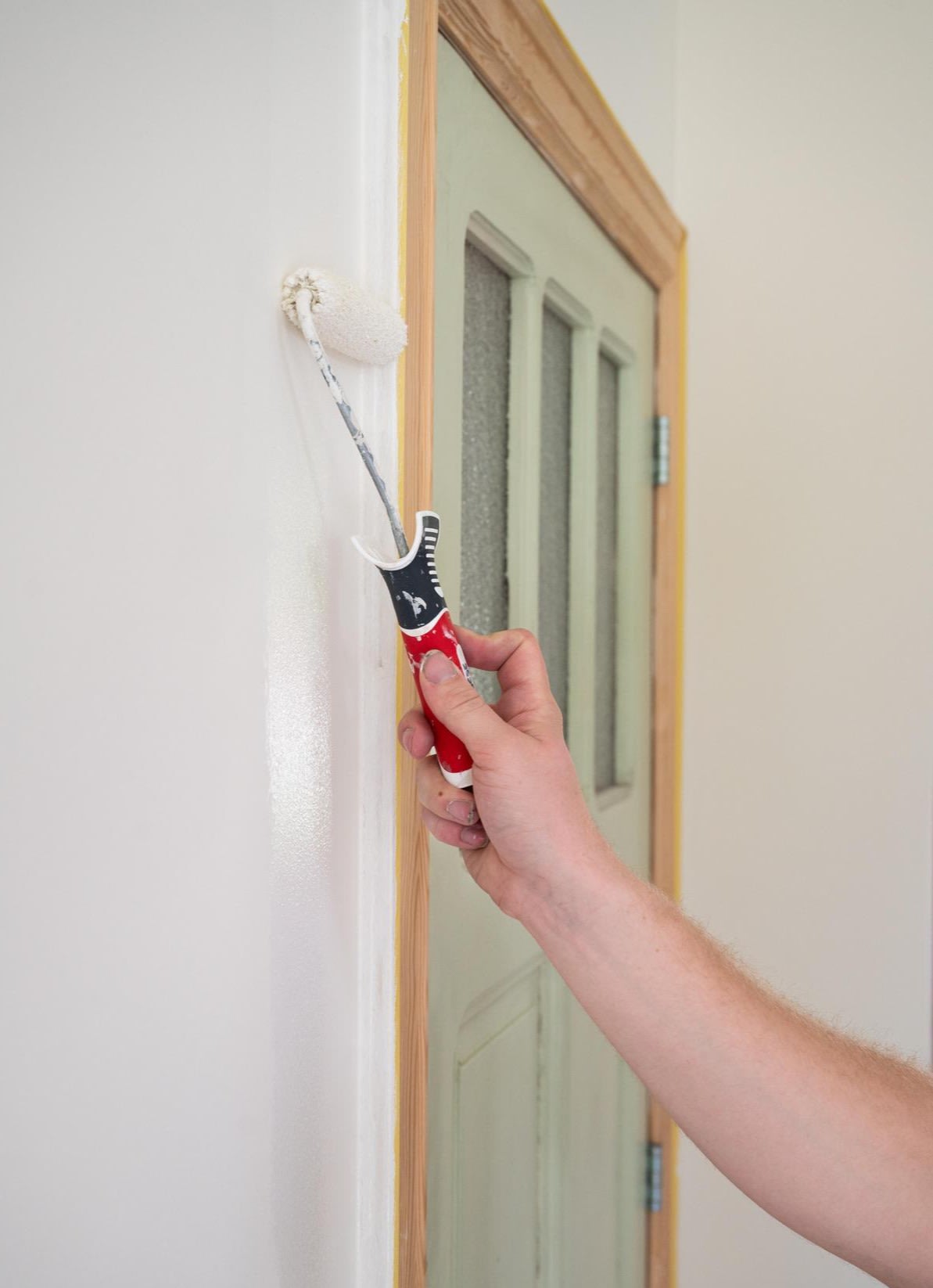 Quelqu'un peint un mur blanc à l'aide d'un petit rouleau muni d'un manche rouge et noir, à côté d'une porte vert clair ornée d'une moulure décorative en bois et de trois charnières métalliques.
