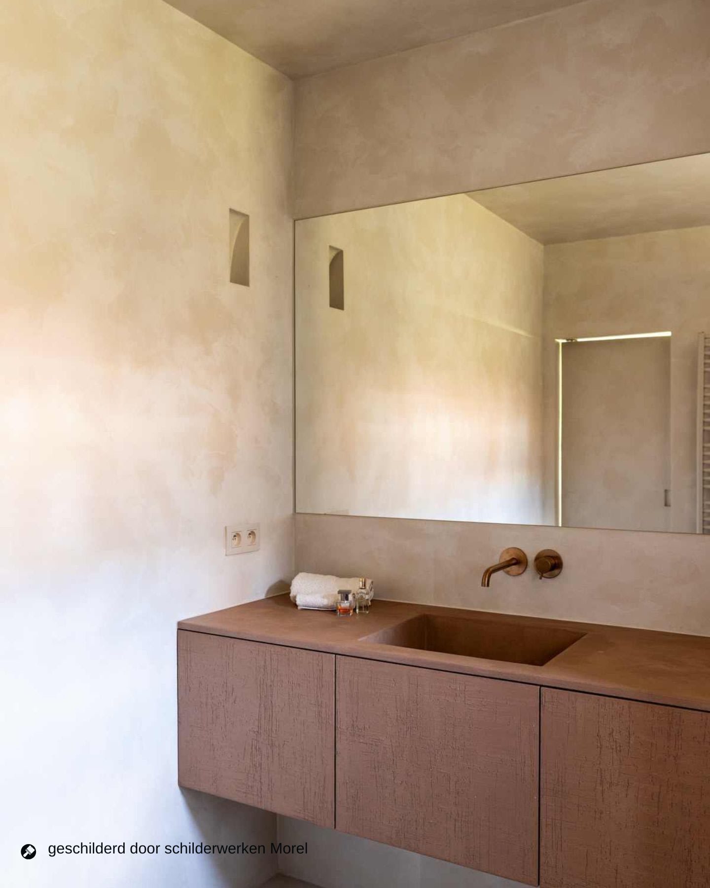 Minimalistische badkamer met beige gestructureerde muren, een grote spiegel, een aan de muur gemonteerde kraan en een bruin zwevend badmeubel met een ingebouwde wastafel en kleine toiletartikelen op het aanrecht.