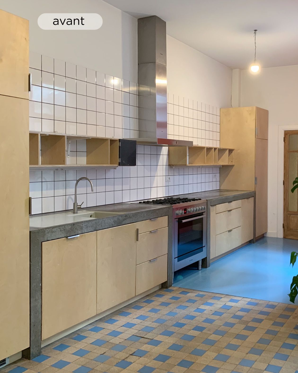 Cuisine avec placards en bois, plan de travail en béton et hotte en inox. Le sol est composé de carreaux carrés et de béton, et le dosseret est recouvert de petits carreaux blancs.
