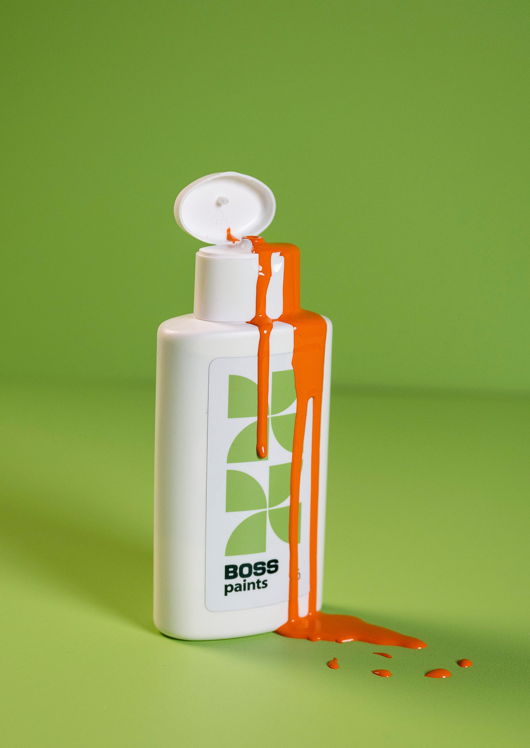 Een witte plastic fles met het label "BOSS paints" staat op een groen oppervlak, met de dop open. Er stroomt feloranje verf uit die langs de fles naar beneden druppelt en op het oppervlak eronder terechtkomt.