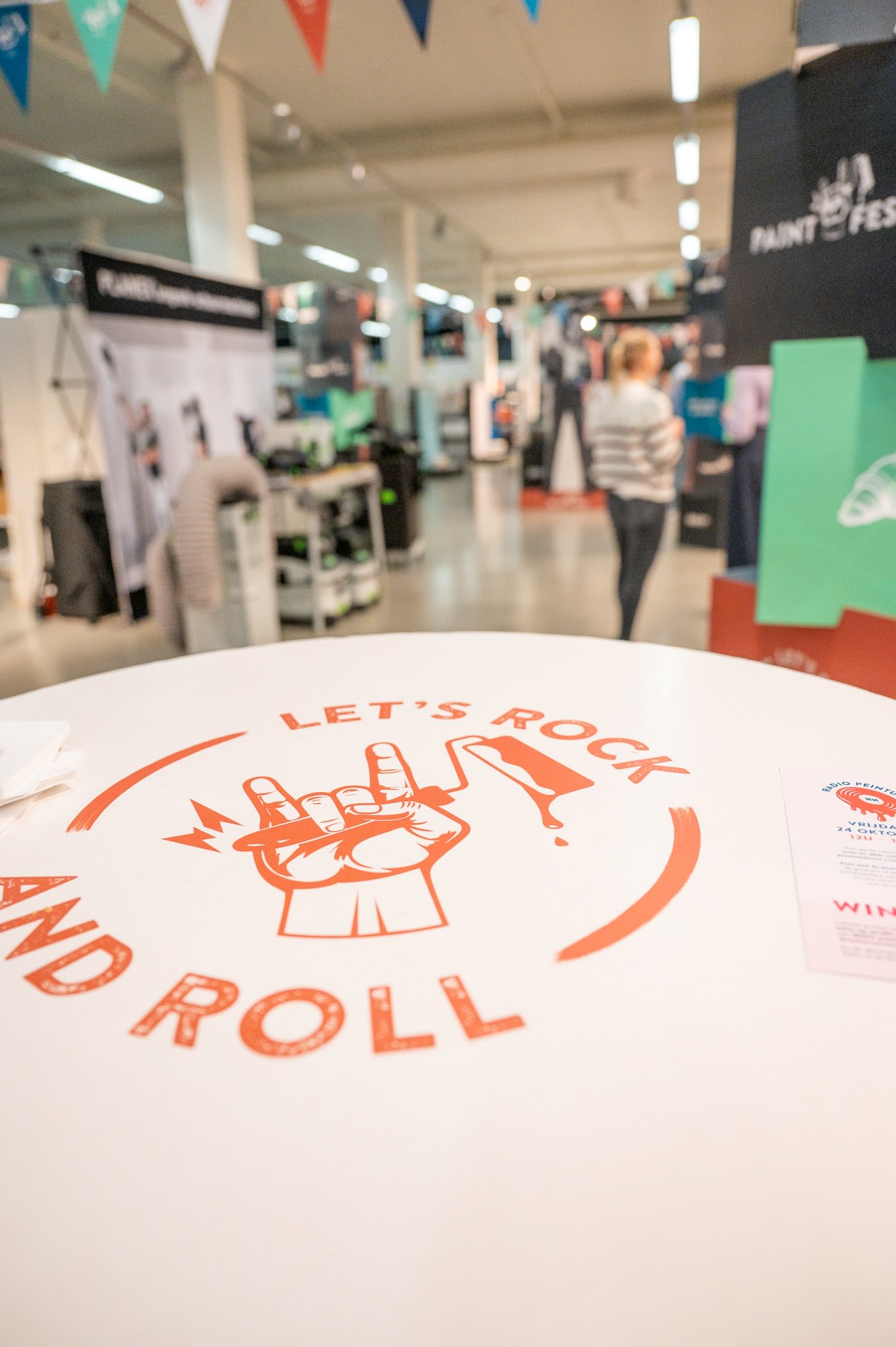 Een ronde witte tafel met "LET'S ROCK AND ROLL" en een handgebaar erop gedrukt; mensen en kraampjes zijn zichtbaar in de onscherpe achtergrond.