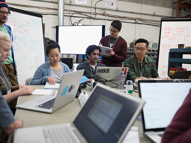 Grupo de siete personas trabajando juntas en el mismo escritorio, en diferentes computadoras.